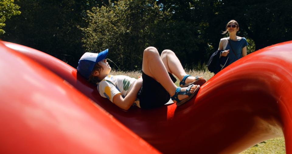 Jonge in zomerse outfit ligt op het rode kunstwerk Play Sculpture van Isamu Noguchi in het kunstpark van het Middelheimmuseum.